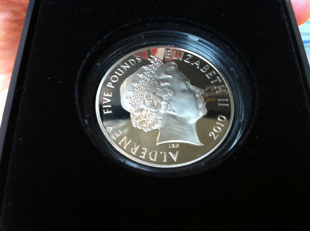 O verso da moeda - "God Save the Queen". Aos curiosos, "Alderney" é a ilha que está mais ao norte do conglomerado conhecido como "Channel Islands", da coroa britânica. A população local é de 2.400 habitantes (Wikipedia)